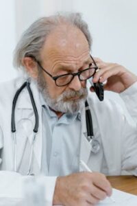 A senior doctor with eyeglasses talks on the phone and writes notes at a desk.