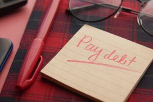 Close-up of a note reading 'Pay debt' next to a red pen on a plaid fabric, emphasizing financial reminders.